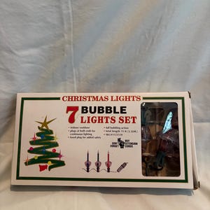 May include: A boxed set of 7 bubble Christmas lights with a total length of 11 ft. The white packaging features a green border, a graphic of a decorated Christmas tree, and text including "7 BUBBLE LIGHTS SET," "indoor/outdoor," and "fused plug for added safety."