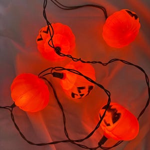 May include: A string of five glowing orange pumpkin-shaped lights connected by a thin black wire. Each pumpkin features a carved jack-o'-lantern face with black triangular eyes and a jagged mouth, emitting a warm, bright orange light.