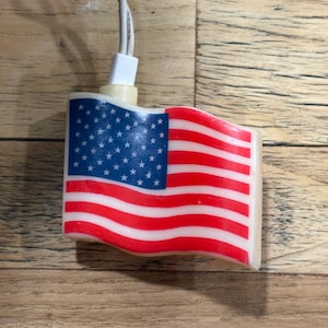 May include: A small, decorative light shaped like the American flag. The flag features a blue field with white stars and red and white stripes. A white cord and plug extend from the top of the flag.