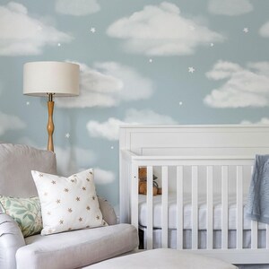 May include: A nursery room featuring light blue wallpaper with a white cloud and star pattern, a white wooden crib, a light gray upholstered armchair with a matching ottoman, a floor lamp with a beige shade, and decorative throw pillows.