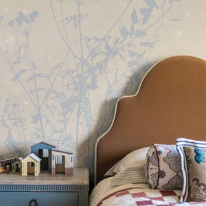 May include: A bedroom scene featuring a brown headboard with a scalloped edge and a patterned quilt. A blue nightstand holds miniature wooden houses. The wall is adorned with a floral pattern in soft blue and beige tones, creating a cozy atmosphere.