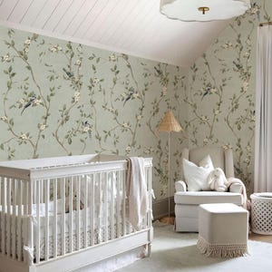 May include: A nursery room featuring light green wallpaper with a repeating pattern of brown branches, green leaves, white flowers, and small birds. The room includes a white wooden cot, a beige upholstered armchair, a matching ottoman, and a lamp.