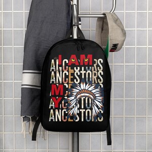 May include: Black backpack with the text "I AM MY ANCESTORS" in a distressed font. The design includes a Native American headdress in shades of blue, orange, and white. The backpack is hanging on a metal hook.