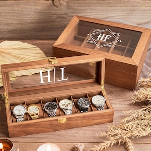 May include: Two wooden watch display boxes with glass lids, one open and one closed. The open box displays six watches with silver and gold bands. The lids feature initials. The boxes sit on a wooden surface.