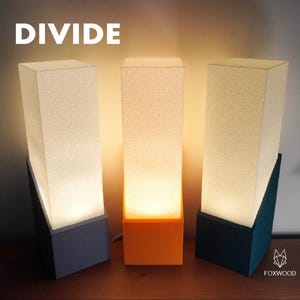 Puede incluir: Tres lámparas modernas con pantallas rectangulares y bases anguladas. Las pantallas son blancas y texturizadas, y las bases son en tonos azules y naranjas. La palabra "DIVIDE" está impresa en blanco en la parte superior. Las lámparas están sobre una superficie de madera.