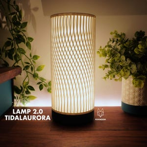 Puede incluir: Una lámpara cilíndrica con una pantalla crema estampada y una base negra. La lámpara está encendida, emitiendo un brillo cálido. El texto "LAMP 2.0 TIDALAUROA" es visible. Un elemento decorativo para el hogar.