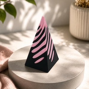 Op de afbeelding: Een zwart en roze geometrisch piramidevormig object met een zebra streep ontwerp. Het object wordt weergegeven op een grijs cirkelvormig platform. De piramide is gemaakt van een zwart materiaal met roze strepen. De achtergrond bevat een plant en een vaas.