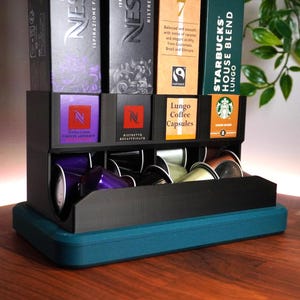 May include: A black coffee pod holder with a teal base, holding various coffee pod boxes and individual pods. The boxes include Nespresso and Starbucks brands. The holder is on a wooden surface.