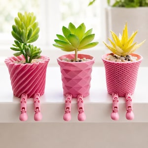 May include: Three pink decorative planters with faux succulents. The planters have unique textures and are supported by pink legs. The succulents are green and yellow, and the planters are filled with small pebbles. A creative and colorful home decor item.