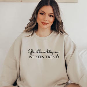 Könnte beinhalten: Ein beiges Sweatshirt mit dem Text "Gleichberechtigung IST KEIN TREND" in schwarzer Schrift. Das Sweatshirt hat eine neutrale Farbe und einen Rundhalsausschnitt. Das Bild zeigt eine Person, die das Sweatshirt trägt.