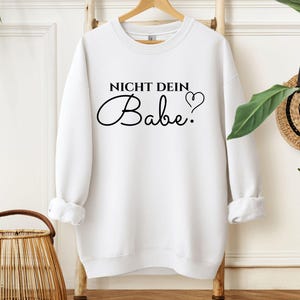May include: White sweatshirt with the text "NICHT DEIN Babe." in black script. A heart symbol follows the word "Babe." The sweatshirt is displayed on a wooden clothing rack. The sleeves are cuffed.
