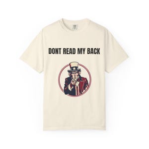 Könnte beinhalten: Cremefarbenes T-Shirt mit einer Grafik von Onkel Sam, der mit dem Finger zeigt, umrandet in Rot und Blau. Der Text "DONT READ MY BACK" ist über der Grafik aufgedruckt. Ein lässiges Kleidungsstück.