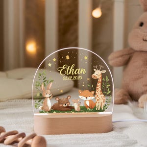 Op de afbeelding: Nachtlampje met een houten basis en een helder acryl paneel. Het paneel toont een bosscène met een konijn, beer, vos en giraffe, samen met de naam "Ethan" en een datum. Het licht geeft een zachte gloed.