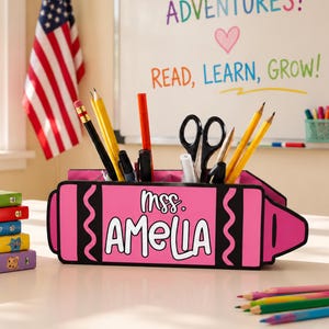 May include: A pink crayon-shaped desk organizer filled with pencils, pens, and scissors. The organizer has the name "Amelia" printed on it. An American flag hangs in the background, along with a whiteboard with the words "Read, Learn, Grow!"
