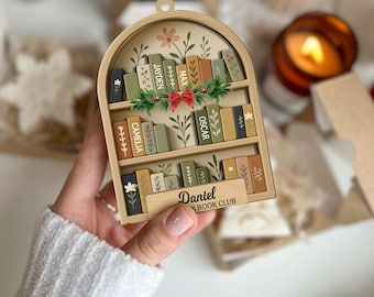 May include: A wooden bookshelf ornament with a decorative arch top. The ornament features miniature books with names like "Jayden" and "Oscar." A red bow and greenery adorn the middle shelf. The bottom shelf reads "Daniel Book Club."