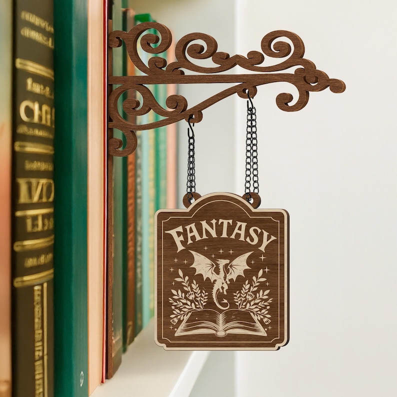 Puede incluir: Un letrero de madera con la palabra "FANTASY" en una fuente decorativa, colgado de un soporte decorativo. El letrero presenta una ilustraci&oacute;n de un drag&oacute;n sobre un libro abierto. El soporte est&aacute; sujeto a una estanter&iacute;a con libros.