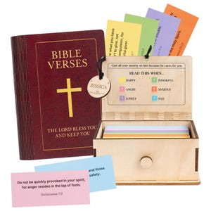Puede incluir: Un libro de la Biblia de color burdeos con letras doradas y una cruz dorada, junto a una caja de madera con tarjetas de colores. El libro dice "BIBLE VERSES" y "THE LORD BLESS YOU AND KEEP YOU". La caja tiene tarjetas con versículos bíblicos.