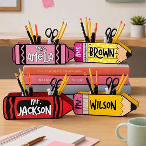 May include: Four personalized pencil and crayon-shaped desk organizers. The pink crayon-shaped organizer reads "mss. AMELIA". The yellow pencil-shaped organizer reads "Mrs. BROWN". The red crayon-shaped organizer reads "Mr. JACKSON". The yellow pencil-shaped organizer reads "Mr. WILSON".