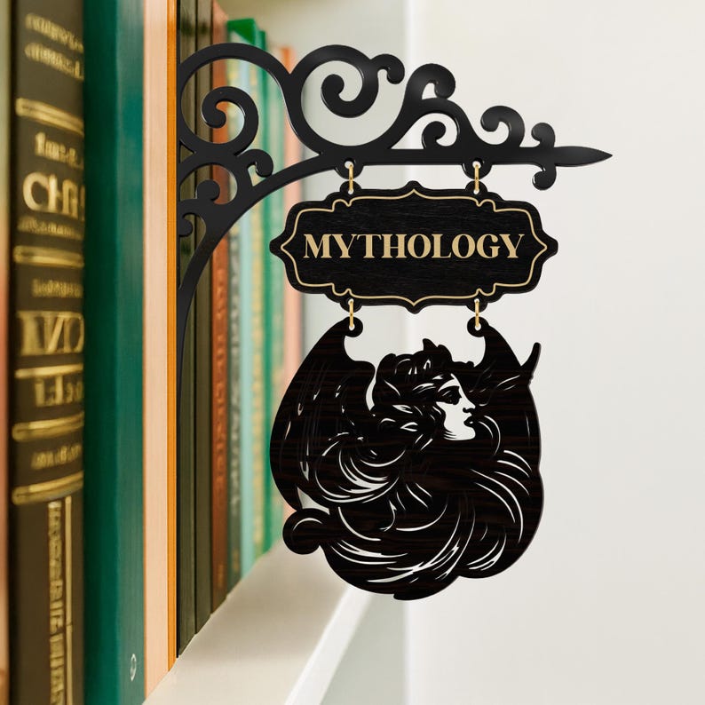 以下が含まれることがあります： 金色の文字で「MYTHOLOGY」と書かれた黒い金属製の看板。看板は、翼のある人物のシルエットが特徴的な装飾的なデザインです。看板は本棚に掛けられています。