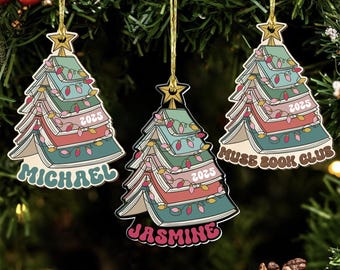 Könnte beinhalten: Drei Weihnachtsbaumschmuckstücke in Form von Bücherstapeln, mit einem Stern oben. Jedes Ornament trägt den Namen "Michael", "Jasmine" und "Muse Book Club". Sie sind mit bunten Lichtern und dem Jahr "2025" verziert.