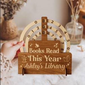 Puede incluir: Un rastreador de libros de madera con indicadores numéricos arqueados y una base que dice "Books Read This Year Ashley's Library". Los números están grabados en la madera, con elementos decorativos como una vela, pájaros y un libro.