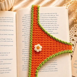 May include: A triangular crochet bookmark featuring a vibrant orange center with a textured green border. A small white flower with a yellow center is attached to the middle of the orange section. The bookmark is placed vertically inside an open book.