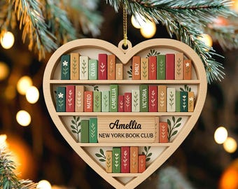 Könnte beinhalten: Herzförmiger Holzschmuck mit Bücherregal-Design. Der Schmuck hat zwei Regale mit bunten, Miniaturbüchern. Die Bücher sind mit Namen wie "Olivia" und "Mateo" beschriftet. Der Schmuck enthält auch den Text "Amellia NEW YORK BOOK CLUB."
