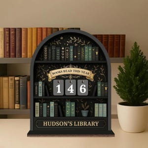 May include: A black wooden bookshelf-shaped display with the text "BOOKS READ THIS YEAR" and the number "146". The display also has the text "HUDSON'S LIBRARY" and decorative miniature books. A small potted evergreen tree is to the right.