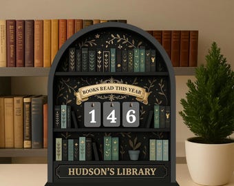 May include: A black wooden bookshelf-shaped display with the text "BOOKS READ THIS YEAR" and the number "146". The display also has the text "HUDSON'S LIBRARY" and decorative miniature books. A small potted evergreen tree is to the right.