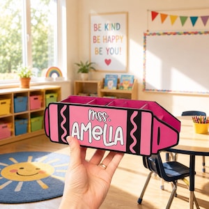May include: A pink crayon-shaped desk organizer with the name "mrs. AMELIA" in white lettering. The organizer has multiple compartments and is held in a classroom setting. The background includes a whiteboard, colorful decor, and a "Be Kind" sign.