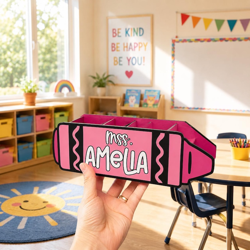 May include: A pink crayon-shaped desk organizer with the name "mrs. AMELIA" in white lettering. The organizer has multiple compartments and is held in a classroom setting. The background includes a whiteboard, colorful decor, and a "Be Kind" sign.