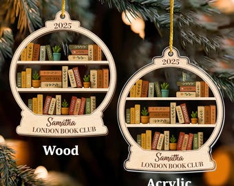 Könnte beinhalten: Zwei Weihnachtsbaumschmuck aus Holz und Acryl in Form eines Bücherregals, mit dem Text "2025" oben. Die Ornamente zeigen Miniaturbücher, Pflanzen und den Text "Samatha LONDON BOOK CLUB". Die Wörter "Wood" und "Acrylic" sind ebenfalls abgebildet.
