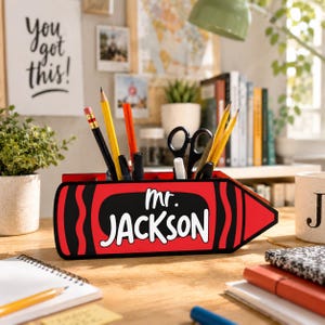 May include: A red crayon-shaped desk organizer with the name "Mr. Jackson" in white. The organizer holds pencils, pens, and scissors. A motivational poster that says "You got this!" is in the background.