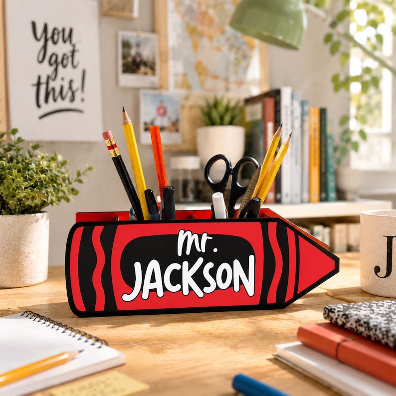 May include: A red crayon-shaped desk organizer with the name "Mr. Jackson" in white. The organizer holds pencils, pens, and scissors. A motivational poster that says "You got this!" is in the background.