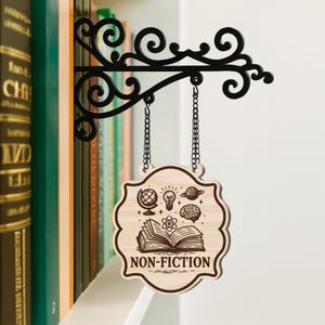 以下が含まれることがあります： 本棚用の装飾的な木製サインで、「NON-FICTION」と書かれています。サインは、装飾的な渦巻き模様のある黒い金属製のブラケットからチェーンで吊り下げられています。サインには、地球儀、電球、惑星、脳、開いた本のイラストが描かれています。