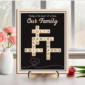 May include: A framed family sign with the words "Family is the heart of a home" and "Our Family" in elegant script. The black background features a crossword puzzle design with family names spelled out in wooden letter tiles. A heart graphic is at the bottom.