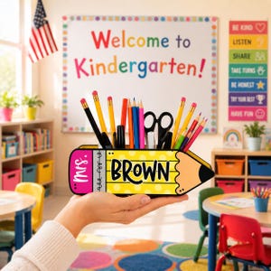 May include: A yellow pencil-shaped desk organizer with the name "BROWN" in black letters. The organizer holds pencils, pens, and scissors. The word "Mrs." is on the pink eraser end. The background is a classroom setting.