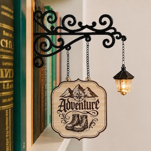 May include: A black metal wall-mounted bracket with scrollwork, featuring a hanging wooden sign with the word Adventure, a mountain and compass graphic, and a pair of boots. A small black lantern with a warm glowing light hangs from the bracket.