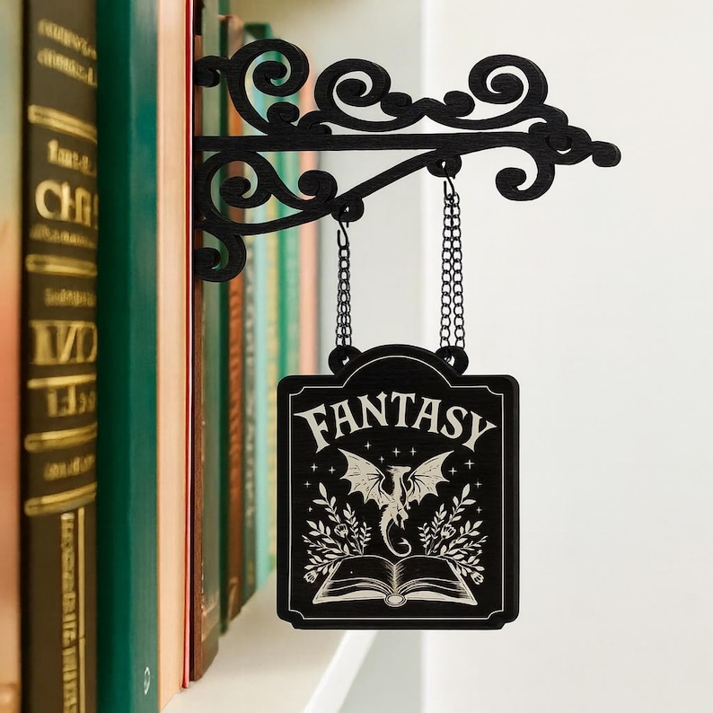 K&ouml;nnte beinhalten: Schwarzes Metallschild "Fantasy", das an einer dekorativen Halterung h&auml;ngt. Das Schild zeigt eine Drachenillustration &uuml;ber einem offenen Buch mit dem Wort "FANTASY". Die Halterung ist an einem Regal mit B&uuml;chern befestigt.
