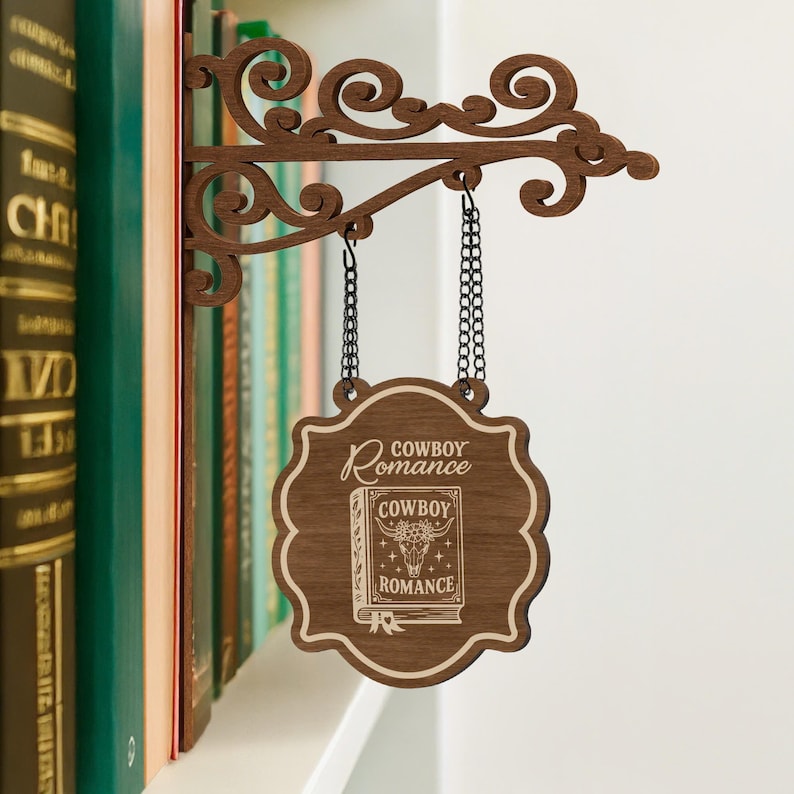 Puede incluir: Un letrero de estanter&iacute;a de madera con las palabras "Cowboy Romance" y una ilustraci&oacute;n de libro. El letrero es marr&oacute;n con un soporte decorativo y cadenas colgantes. El letrero cuelga frente a una estanter&iacute;a.