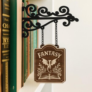 May include: A black metal bookshelf sign with a decorative scroll design, hanging from a chain. The wooden sign reads "FANTASY" with a dragon and open book illustration. The sign is displayed next to a row of books.