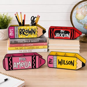 May include: Four colorful pencil and crayon-shaped desk organizers. One yellow pencil organizer reads "BROWN" and another reads "WILSON". A red crayon organizer reads "JACKSON" and a pink crayon organizer reads "AMELIA".