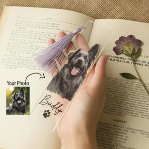 May include: A clear acrylic bookmark featuring a photo of a black and gray dog with its tongue out. The bookmark has the name "Buddy" and a paw print. A light purple tassel hangs from the top. The words "Your Photo" are also visible.