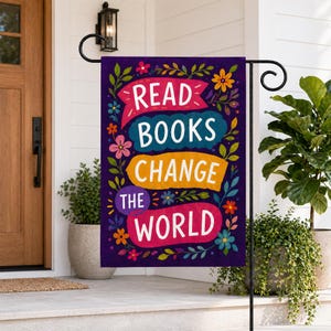 May include: A purple garden flag with the words "READ BOOKS CHANGE THE WORLD" in white, teal, and yellow banners. The flag is decorated with colorful flowers and green leaves. The flag is hanging on a black metal stand.