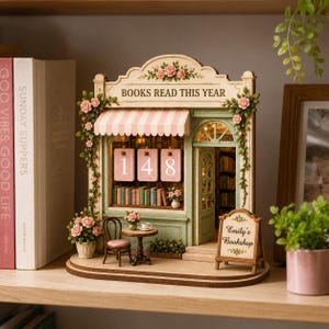 May include: A miniature wooden bookstore model with a pink and white striped awning, featuring the text "BOOKS READ THIS YEAR" and "Emily's Bookshop." The model includes a small table, chair, and a sign displaying the number "148."