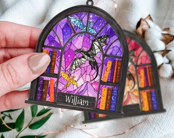 May include: A wooden ornament shaped like a stained-glass window, featuring a fantasy scene with dragons, books, and the name "William". The window design incorporates shades of purple, pink, and blue, with a black frame.