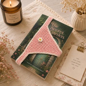 May include: A pink and white crocheted bookmark with a daisy accent rests on a copy of "The Secret Garden" by Frances Hodgson Burnett. A lit candle and a small vase with flowers are also in the image. The candle reads "Reading is my Happy Hour."