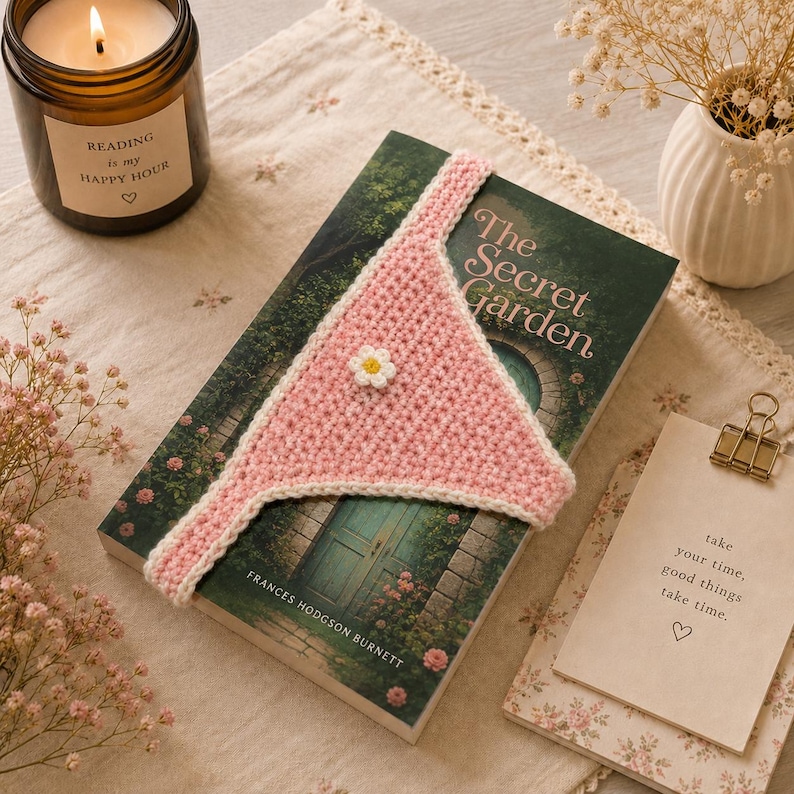 May include: A pink and white crocheted bookmark with a daisy accent rests on a copy of "The Secret Garden" by Frances Hodgson Burnett. A lit candle and a small vase with flowers are also in the image. The candle reads "Reading is my Happy Hour."