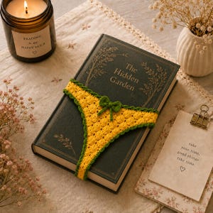 Puede incluir: Un libro de tapa dura verde oscuro titulado The Hidden Garden con una banda elástica de ganchillo amarilla que presenta un pequeño lazo verde alrededor de la cubierta. La banda amarilla tiene un patrón circular texturizado y un borde de ganchillo verde.