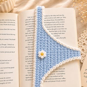 May include: A handmade, light blue crochet bookmark with a white scalloped edge and a small white and yellow daisy. The bookmark is placed in an open book, adding a touch of handmade charm to reading.